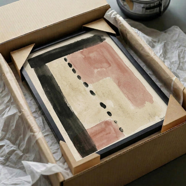 May include: Framed abstract art print in cardboard shipping box with protective wrapping. Artwork features soft terracotta pink and sage green brushstrokes with scattered black dots on cream background. Black frame sits among white tissue paper and wooden dowels used for safe packaging and transport.