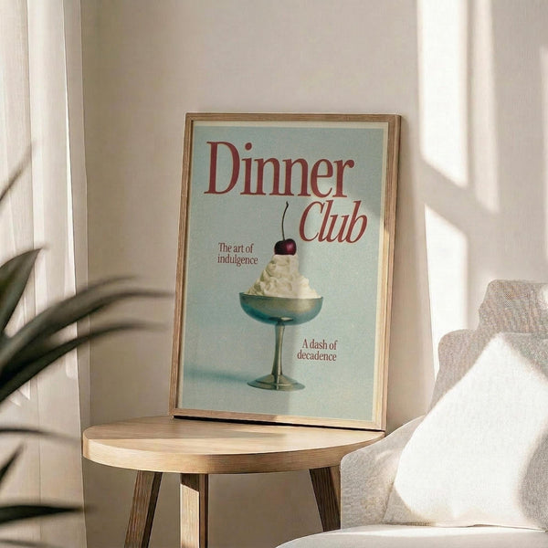 May include: Framed vintage-style poster with light blue background featuring red "Dinner Club" text and illustration of whipped cream dessert in turquoise cocktail glass with cherry on top. Text reads "The art of indulgence" and "A dash of decadence." Natural wood frame sits on round wooden side table next to beige upholstered chair with throw pillow. Soft natural lighting creates shadows on cream wall with plant leaves visible in corner.