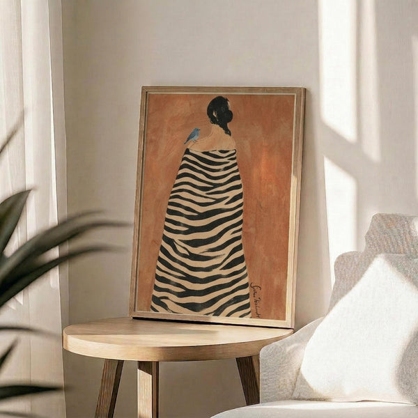 May include: Framed artwork displayed on a round wooden side table next to a beige upholstered chair. The print features a figure in profile wearing a black and white striped zebra-patterned cloak against a warm orange background, with a small blue bird perched on their shoulder. The piece is in a thin gold frame and shows earthy, painterly textures. Soft natural lighting creates shadows across the neutral-toned room setting with a plant visible in the corner.