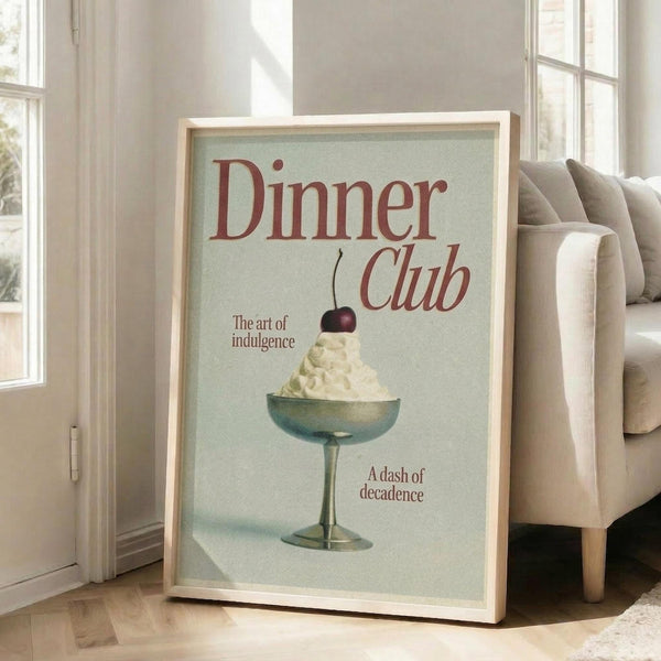 May include: Framed vintage-style poster with pale mint green background displaying red text reading "Dinner Club" and "The art of indulgence" alongside an illustration of a coupe glass filled with white whipped cream topped with a dark cherry. The poster reads "A dash of decadence" at bottom. Light wood frame sits on hardwood floor against white walls near beige upholstered chair and tall windows with natural lighting.