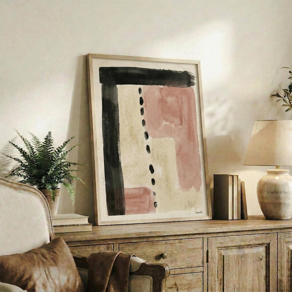 May include: Abstract watercolor art print in light wood frame displayed on rustic wooden sideboard alongside fern plant, ceramic vases, and stacked books. Artwork features muted terracotta pink, cream, and charcoal black brushstrokes with dotted vertical line details. Warm, minimalist home decor setting with neutral tones and natural textures.