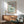 May include: Framed vintage-style poster displaying a mint green dessert glass with whipped cream and cherry, surrounded by red text reading "Dinner Club" and "The art of indulgence" and "A dash of decadence." Shown in modern living room with mid-century wooden credenza, sage green upholstered chair with wooden arms, white ceramic table lamp, and natural sunlight casting shadows on white wall.