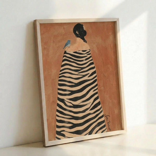May include: Framed art print leaning against white wall showing woman from behind wearing zebra-striped cloak or dress, with dark braided hair and small blue bird perched on shoulder, painted on warm peachy-orange background, displayed in light wood frame on white surface
