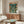 May include: Vintage-style Neon Genesis Evangelion poster displayed above a mid-century modern walnut credenza in a minimalist living room. The poster features a large green mecha robot towering over a small figure on a city street, with Japanese text at the top. Room includes a white curved chair, oval glass coffee table, and modern lighting fixtures against light walls.