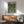 May include: Framed Neon Genesis Evangelion poster displayed above a wooden sideboard in a mid-century modern living room. The vintage-style poster shows a young pilot character standing in a futuristic city street with a large mecha towering behind him in green and teal tones. Japanese text appears at the top. The room features a teal upholstered armchair, round coffee table, and white ceramic lamp with natural sunlight streaming across white walls.