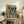 May include: Framed anime poster featuring a young male character standing in a neon-lit urban street with a large mecha robot towering behind him, displayed on a rustic wooden sideboard next to a fern plant, ceramic vase, and books in a cozy living room setting with beige armchair visible