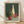 May include: Framed still life painting leaning against white wall on wooden floor. Artwork shows three ceramic vases in green, cream, and terracotta colors alongside a red apple, set against dark green background. Natural wood frame with cream matting. Soft natural lighting creates diagonal shadows across the scene.