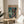 May include: Framed anime poster displayed on wooden round side table next to beige upholstered chair. Poster features Japanese text at top, depicts young man in white pilot suit standing beside large orange and purple mechanical robot in rain. Artwork has retro anime illustration style with muted teal and orange color palette. Natural wooden frame matches table. Soft window lighting creates shadows across white wall and plant leaves visible at left edge.