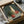 May include: Framed still life painting being unpacked from shipping box with protective tissue paper. Artwork shows green ceramic vase, tall beige bottle, red apple, and orange persimmon against sage green background. Black frame with white mat, surrounded by cardboard packaging materials on white surface.
