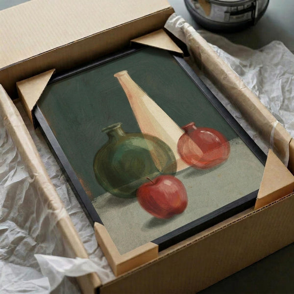 May include: Framed still life painting being unpacked from shipping box with protective tissue paper. Artwork shows green ceramic vase, tall beige bottle, red apple, and orange persimmon against sage green background. Black frame with white mat, surrounded by cardboard packaging materials on white surface.