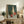 May include: Still life painting in light wood frame showing green glass bottle, tall cream vase, small red glass vessel, and red apple arranged on neutral surface against teal background. Artwork displayed on rustic wooden sideboard with fern plant, books, ceramic pottery, and brown leather chair visible in cozy living room setting with white walls.