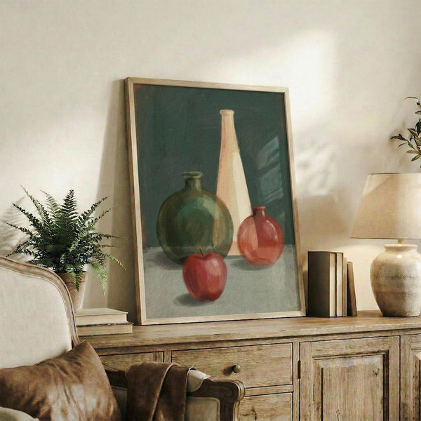 May include: Still life painting in light wood frame showing green glass bottle, tall cream vase, small red glass vessel, and red apple arranged on neutral surface against teal background. Artwork displayed on rustic wooden sideboard with fern plant, books, ceramic pottery, and brown leather chair visible in cozy living room setting with white walls.