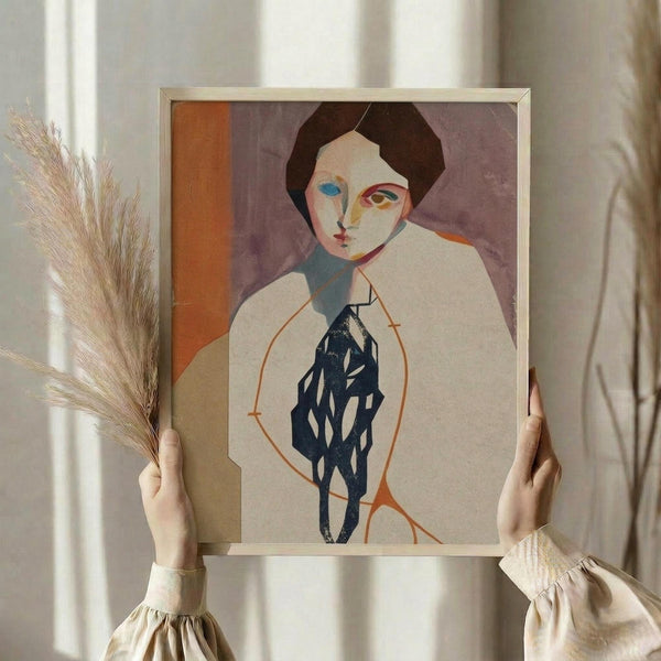 May include: Hands holding a framed modernist portrait artwork featuring a figure with dark hair, colorful geometric face painting in blues and oranges, and a dark patterned garment against a cream background. One hand also holds dried wheat stalks. The frame appears light wood-toned against a neutral wall with soft natural lighting.