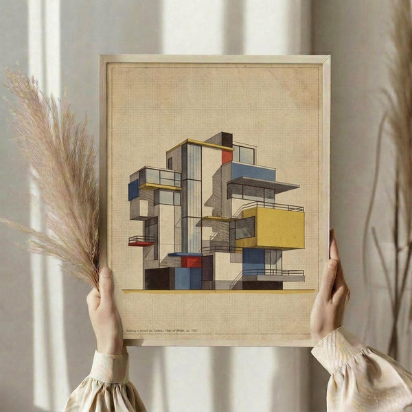 May include: Person holding framed modernist architectural print featuring geometric building design with colorful blocks in red, blue, yellow, and gray. Light wood frame against textured beige background. Dried pampas grass visible to left. Person wearing cream-colored top with gathered sleeves in soft natural lighting near window.