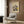 May include: Framed architectural artwork showing a modernist building design with geometric colored blocks in yellow, blue, red and white on cream background, displayed on wood paneled wall above beige upholstered armchair with patterned pillow, brass floor lamp and side table with open book nearby