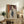 May include: Framed modernist portrait artwork displayed on rustic wooden sideboard in neutral-toned living room. The stylized painting features a figure with dark hair in white clothing against purple and orange geometric background. Warm lighting illuminates the wood furniture, decorative pottery, books, and fern plant creating a cozy interior scene.