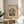 May include: Framed architectural artwork featuring a modernist building design with geometric shapes in yellow, blue, red, and gray colors, displayed on a light wood round side table next to a beige upholstered chair. Natural sunlight streams across white walls, with green plant leaves visible in the corner. The artwork shows an abstract, Bauhaus-style building composition on cream background within a thin wooden frame.