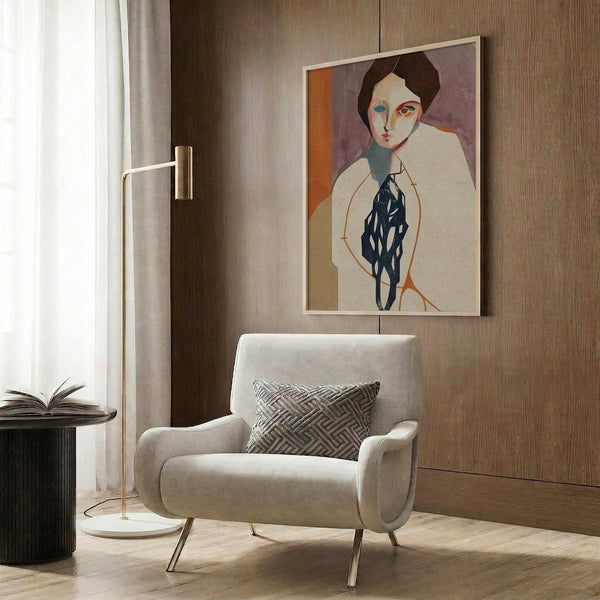 May include: A framed modernist portrait print featuring a woman with brown hair and stylized facial features in cream, orange, and navy blue tones, displayed on wood paneled walls above a beige upholstered armchair with geometric patterned pillow, alongside a brass floor lamp and dark round side table with open book.