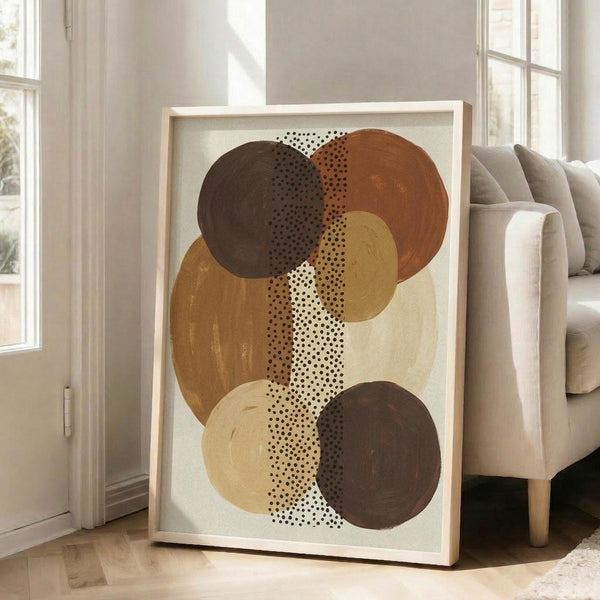 May include: Abstract art print in light wood frame leaning against white wall next to beige sofa. Artwork features overlapping circles in earthy brown, rust, mustard yellow, and cream tones with black dotted pattern running vertically through center. Modern minimalist living room setting with bright natural light from windows.