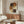 May include: Abstract poster with overlapping circles in earthy brown, rust, mustard, and cream tones connected by dotted vertical line, displayed in thin wooden frame above walnut credenza. Modern living room setting with white curved chair, glass coffee table, and warm natural lighting creating shadows on neutral walls.