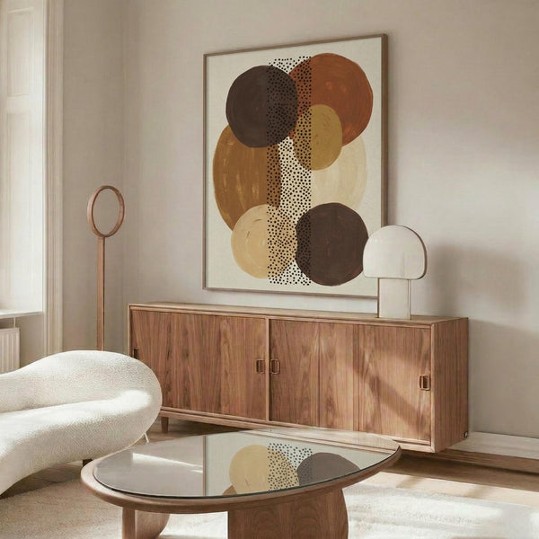 May include: Abstract poster with overlapping circles in earthy brown, rust, mustard, and cream tones connected by dotted vertical line, displayed in thin wooden frame above walnut credenza. Modern living room setting with white curved chair, glass coffee table, and warm natural lighting creating shadows on neutral walls.
