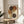 May include: Abstract art print in wooden frame displaying overlapping circles in earthy tones of brown, rust orange, cream and golden yellow with dotted patterns running vertically through center, displayed on round wooden side table next to beige upholstered chair with plant leaves visible in corner, natural sunlight casting shadows on neutral wall