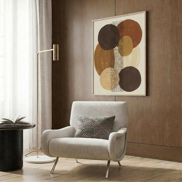 May include: Abstract art print featuring overlapping circles in earthy brown, rust, tan and cream tones with dotted textures, displayed in light wood frame on wooden wall paneling above modern beige armchair with geometric pillow, brass floor lamp and dark side table with open book in contemporary neutral living room setting.