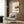 May include: Framed Japanese calligraphy poster with kitchen still life illustrations hanging on wood-paneled wall above beige armchair. Artwork features black vertical text, teapot, bowls, and coffee cup in muted earth tones on cream background. Modern reading nook with geometric pillow, brass floor lamp, and dark round side table with open book.