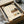 May include: Framed Japanese calligraphy poster with kitchen still life illustration displayed in cardboard shipping box with protective wrapping. Artwork features teapot, dishes, and food items on wooden shelf with Japanese text on cream background. Black frame sits nestled in brown packaging materials with white tissue paper.