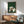 May include: Framed poster with green textured background showing six cats in different colors (white, gray, black, cream) gathered around a wooden table playing a board game. Print displayed above mid-century modern wooden credenza with table lamp, next to sage green upholstered armchair with wooden frame. Natural sunlight creates dramatic shadows on white wall.