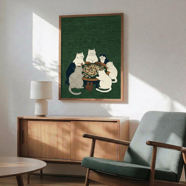 May include: Framed poster with green textured background showing six cats in different colors (white, gray, black, cream) gathered around a wooden table playing a board game. Print displayed above mid-century modern wooden credenza with table lamp, next to sage green upholstered armchair with wooden frame. Natural sunlight creates dramatic shadows on white wall.