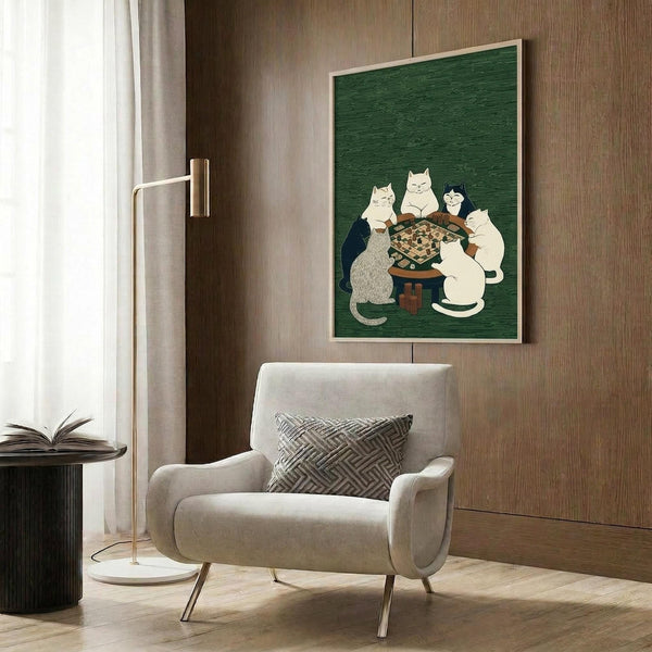 May include: Whimsical poster print featuring five cats gathered around a wooden table playing a board game on deep green textured background with light wood frame, displayed on wood paneled wall above cream upholstered armchair with geometric patterned pillow, beside brass floor lamp and dark side table with open book in modern living room setting