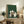 May include: Framed poster with green textured background showing five cats gathered around a wooden table playing chess. Cats include white, gray, black and white varieties sitting on small stools. Print displayed on rustic wooden console with fern plant, books, and ceramic vase in cozy living room setting with beige armchair visible.