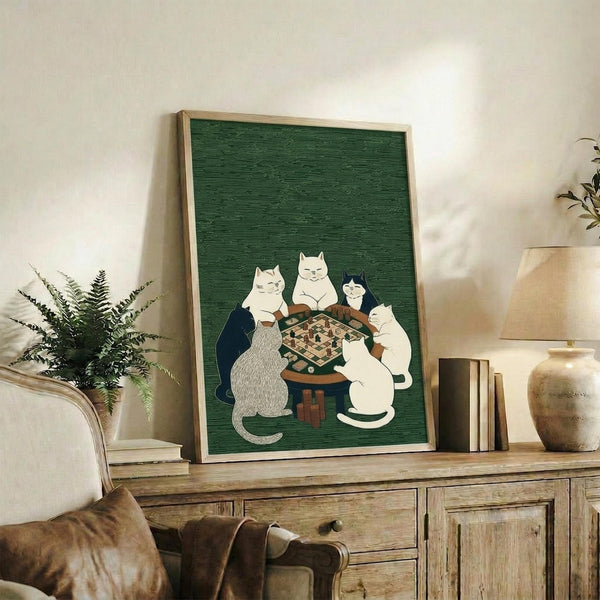 May include: Framed poster with green textured background showing five cats gathered around a wooden table playing chess. Cats include white, gray, black and white varieties sitting on small stools. Print displayed on rustic wooden console with fern plant, books, and ceramic vase in cozy living room setting with beige armchair visible.