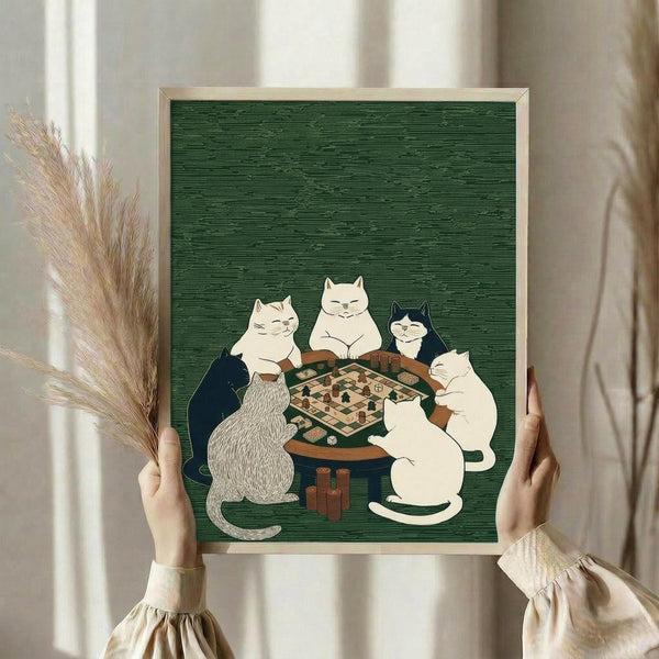 May include: Person holding framed artwork showing five cats gathered around a wooden table playing a board game, with game pieces and dice visible. The illustration has a dark green textured background and features cats in various colors - white, black, and striped. The artwork is displayed in a light wooden frame against white curtains, with dried pampas grass visible to the left.