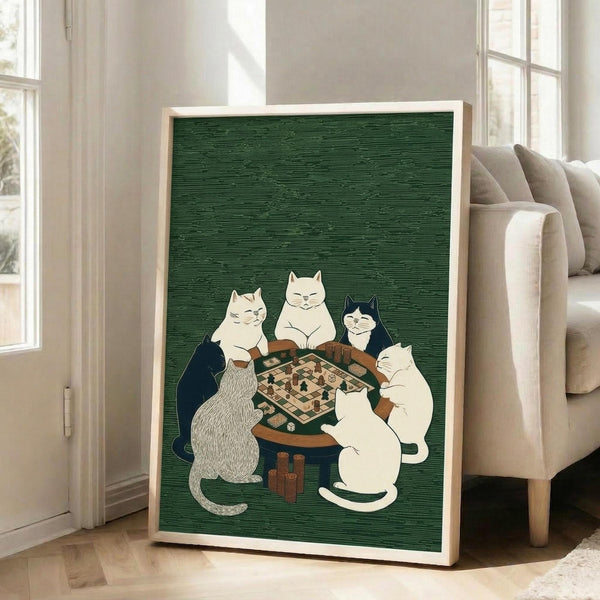 May include: Framed poster with green textured background showing five cats gathered around a wooden table playing a board game. Cats include white, black, gray striped, and black-and-white varieties, all sitting upright. Frame appears light wood, leaning against beige couch in bright living room with white walls and large windows. Natural lighting creates soft shadows on hardwood floors.