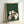 May include: Framed poster print leaning against white wall showing five cats gathered around wooden table playing board game on green textured background. Cats include white, black and white, striped gray, and cream colored felines sitting contentedly around chess or checkers board. Light wooden frame, approximately 12x16 inches, displayed on white surface with soft natural lighting creating gentle shadows.