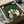May include: Framed artwork with green textured background showing six cats gathered around a wooden table playing a board game. The cats are rendered in cream, white, black, and striped patterns in a folk art illustration style. The piece is being unboxed from cardboard packaging with protective wrapping, positioned on a surface next to what appears to be a dark glass object.