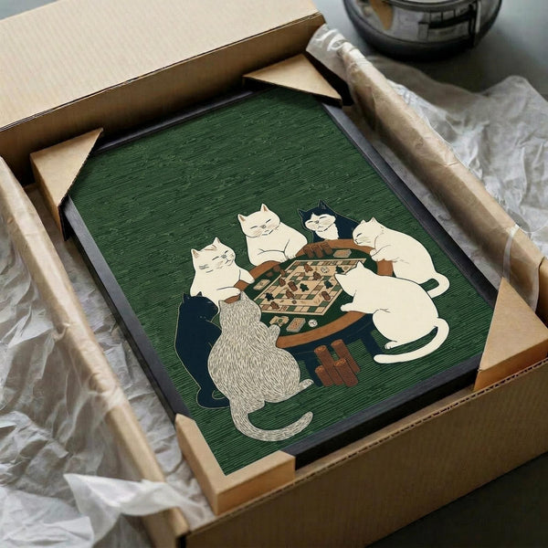 May include: Framed artwork with green textured background showing six cats gathered around a wooden table playing a board game. The cats are rendered in cream, white, black, and striped patterns in a folk art illustration style. The piece is being unboxed from cardboard packaging with protective wrapping, positioned on a surface next to what appears to be a dark glass object.