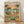 May include: Framed poster print leaning against white wall on light wood floor. Artwork shows two cartoon cats in sunglasses and Hawaiian shirts toasting margaritas at beach table with tacos, guacamole, chips and Mexican food. Tropical beach scene with palm trees and pink sunset sky. Colorful rainbow text reads "FELINE FIESTA" at top. Natural wood frame, vintage-style illustration.