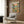 May include: Vintage-style poster showing two cats in tropical shirts and sunglasses sitting at a table with Mexican food including tacos, guacamole, and margaritas. Palm trees and ocean sunset backdrop with "Feline Fiesta" text at top. Poster displayed on wood paneled wall above cream upholstered chair with geometric pillow, gold floor lamp, and side table with open book nearby.