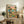 May include: Framed poster print displayed on rustic wooden sideboard showing two cats in sunglasses and Hawaiian shirts at beach table with tacos, margaritas and tropical food. Colorful "FELINE FIESTA" text at top with palm trees and ocean sunset background. Room setting includes potted fern, ceramic vases, books, and brown leather chair.