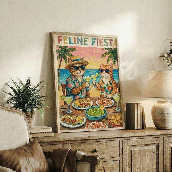 May include: Framed poster print displayed on rustic wooden sideboard showing two cats in sunglasses and Hawaiian shirts at beach table with tacos, margaritas and tropical food. Colorful "FELINE FIESTA" text at top with palm trees and ocean sunset background. Room setting includes potted fern, ceramic vases, books, and brown leather chair.