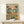 May include: Framed poster print on light wood shelf showing two cats in Hawaiian shirts and sunglasses toasting drinks at beach table with tacos, guacamole, chips and margaritas. Tropical scene with palm trees and ocean backdrop in vintage illustrated style. Small wrapped gift box sits on shelf next to frame against white wall.