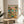 May include: Framed poster artwork featuring two cartoon cats in sunglasses sitting at a beach table with Mexican food and margaritas. The illustration shows palm trees, ocean backdrop, and colorful title reading "Feline Fiesta" at top. Natural wood frame sits on round wooden side table next to beige upholstered chair in bright, airy room with white walls and plant shadows.
