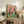 May include: Framed poster with colorful "PENGUIN PARTY" text showing two cartoon penguins at a table with pizza, drinks, and snacks on coral pink background. Wood frame sits on rustic wooden sideboard next to books, pottery vase, and fern plant in cozy living room with beige armchair.