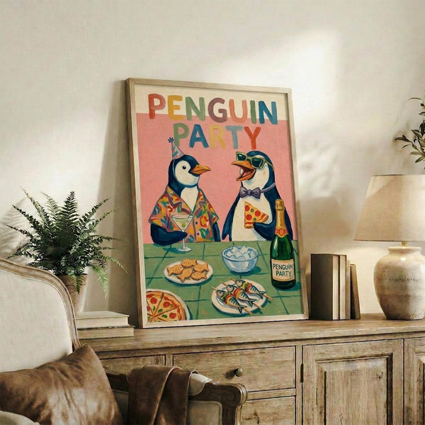 May include: Framed poster with colorful "PENGUIN PARTY" text showing two cartoon penguins at a table with pizza, drinks, and snacks on coral pink background. Wood frame sits on rustic wooden sideboard next to books, pottery vase, and fern plant in cozy living room with beige armchair.