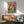 May include: Whimsical framed poster showing two penguins at a colorful party, one wearing a party hat and patterned shirt, the other in a bow tie holding pizza. Table displays pizza slices, fish, drinks and champagne bottle. Rainbow letters spell "PENGUIN PARTY" across coral-pink background. Wood-framed print hangs above mid-century modern wooden credenza with ceramic lamp, positioned next to sage green upholstered armchair in sunlit room with white walls.