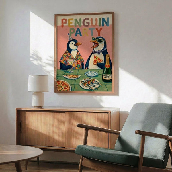 May include: Whimsical framed poster showing two penguins at a colorful party, one wearing a party hat and patterned shirt, the other in a bow tie holding pizza. Table displays pizza slices, fish, drinks and champagne bottle. Rainbow letters spell "PENGUIN PARTY" across coral-pink background. Wood-framed print hangs above mid-century modern wooden credenza with ceramic lamp, positioned next to sage green upholstered armchair in sunlit room with white walls.