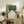 May include: Framed vintage-style bowling alley artwork displayed on rustic wooden sideboard in neutral living room. Print shows green textured bowling lanes with white pins and two red balls, featuring orange Japanese characters. Light wood frame sits among fern plant, ceramic vase, books and cream lamp against white wall.